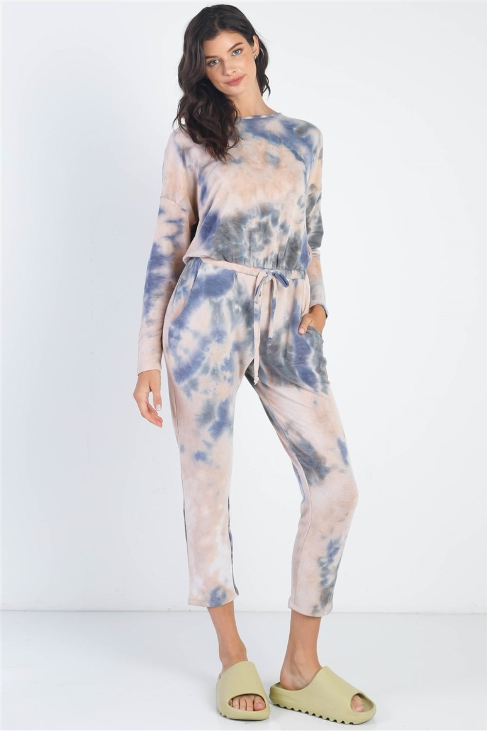 Dressed in a Cherish Apparel Tie-Dye Round Neck Long Sleeve Jumpsuit and green sandals, a person strikes a street-style pose against a plain white background.