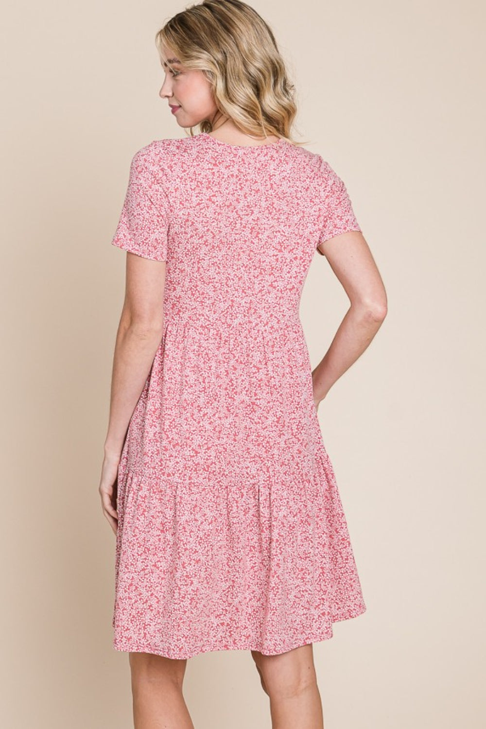 A woman with wavy blonde hair is smiling and holding the side of her BOMBOM Printed Short Sleeve Mini Dress, a lightweight summer dress featuring a pink pattern.