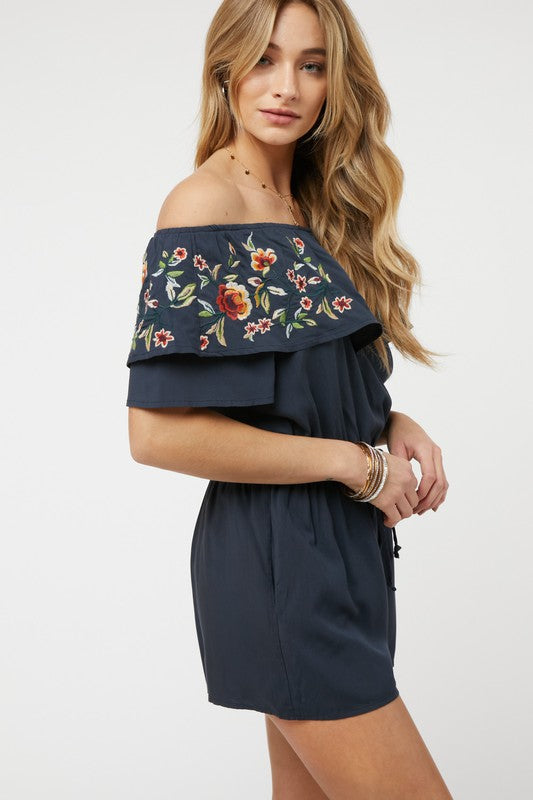 Wearing the stylish Floral Embroidered Off Shoulder Romper, a woman poses gracefully, showcasing its ruffle detail and making it an ideal addition to any summer wardrobe.