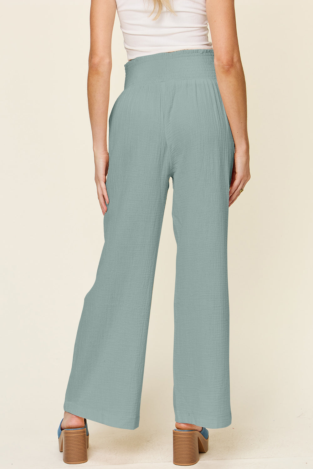 A person wearing Double Take Full Size Texture Smocked Waist Wide Leg Pants in black and a white 100% cotton top is standing with one hand on their hip. They are also wearing blue open-toe sandals.