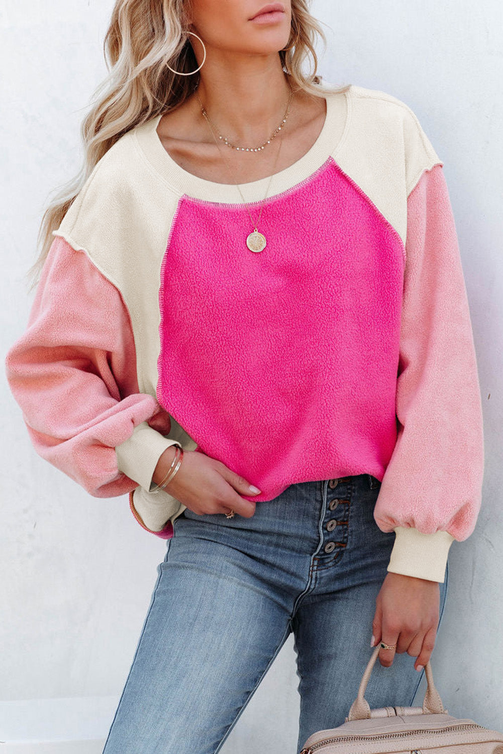 A person with long blonde hair is standing facing away from the camera against a beige background, showcasing the comfortable fleece fabric of the Rose Colorblock Long Sleeve Pullover Fleece Sweatshirt in pink shades and light blue jeans.