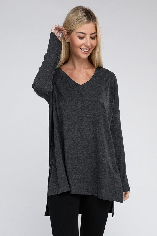 A woman with long, blonde hair is wearing a loose-fitting, brown Dolman Long Sleeve V-Neck Side Slit Hi-Low Hem Top and black pants. She poses with one hand on her hip and a neutral facial expression against a light grey background.