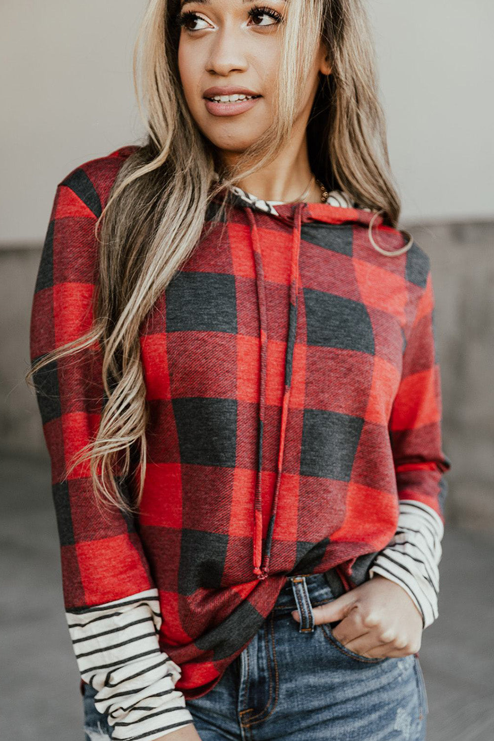 A person stands wearing the Fiery Red Christmas Plaid Striped Patchwork Drawstring Hoodie, characterized by its red and black plaid pattern with striped sleeves. They look to the side with one hand in their pocket, perfectly embodying a casual style.