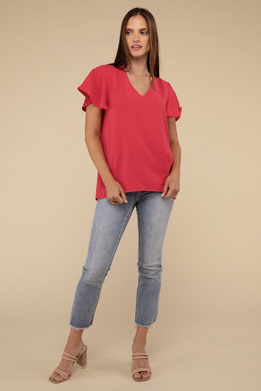 A woman with long straight hair models the Woven Airflow Flutter Sleeve Top in green, matched with light blue jeans, as she poses with her hands in her pockets against a plain beige background.