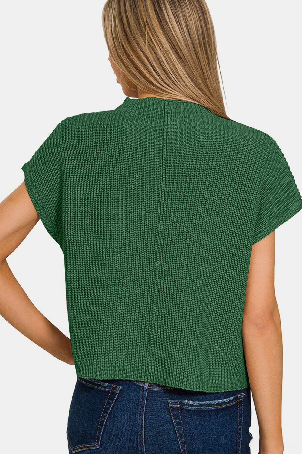 A woman wearing the Zenana Mock Neck Short Sleeve Cropped Sweater in green, featuring a small front pocket and crafted from knit fabric, is standing and smiling. She has long, straight hair and is paired with dark blue jeans.