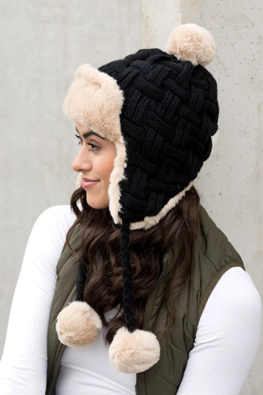 Woman smiling while wearing a brown Cable Weave Pom Trapper Hat with ear flaps and faux fur pom-poms against a neutral background.
