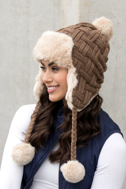 Woman smiling while wearing a brown Cable Weave Pom Trapper Hat with ear flaps and faux fur pom-poms against a neutral background.