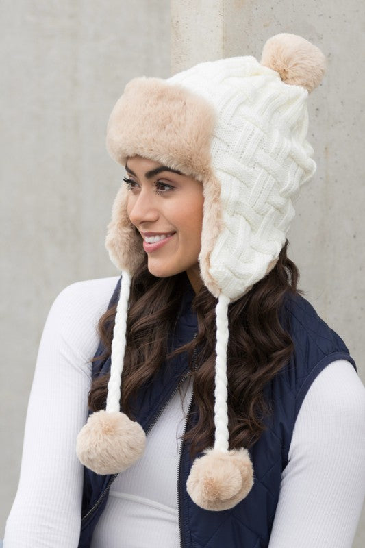 Woman smiling while wearing a brown Cable Weave Pom Trapper Hat with ear flaps and faux fur pom-poms against a neutral background.