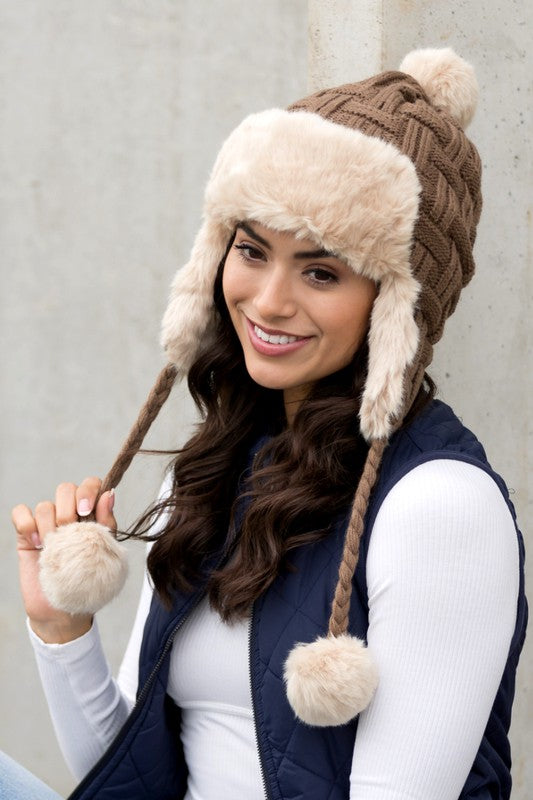 Woman smiling while wearing a brown Cable Weave Pom Trapper Hat with ear flaps and faux fur pom-poms against a neutral background.