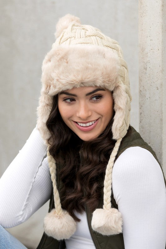 Woman smiling while wearing a brown Cable Weave Pom Trapper Hat with ear flaps and faux fur pom-poms against a neutral background.
