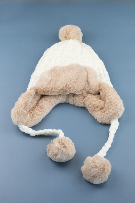 Woman smiling while wearing a brown Cable Weave Pom Trapper Hat with ear flaps and faux fur pom-poms against a neutral background.