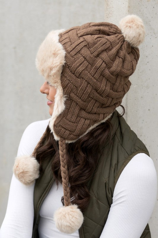 Woman smiling while wearing a brown Cable Weave Pom Trapper Hat with ear flaps and faux fur pom-poms against a neutral background.