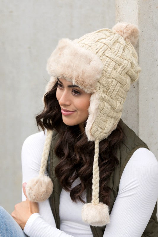 Woman smiling while wearing a brown Cable Weave Pom Trapper Hat with ear flaps and faux fur pom-poms against a neutral background.