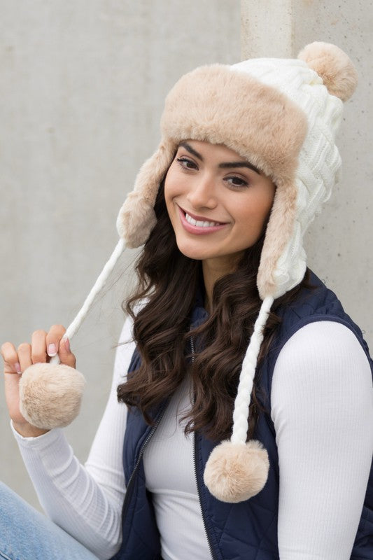 Woman smiling while wearing a brown Cable Weave Pom Trapper Hat with ear flaps and faux fur pom-poms against a neutral background.