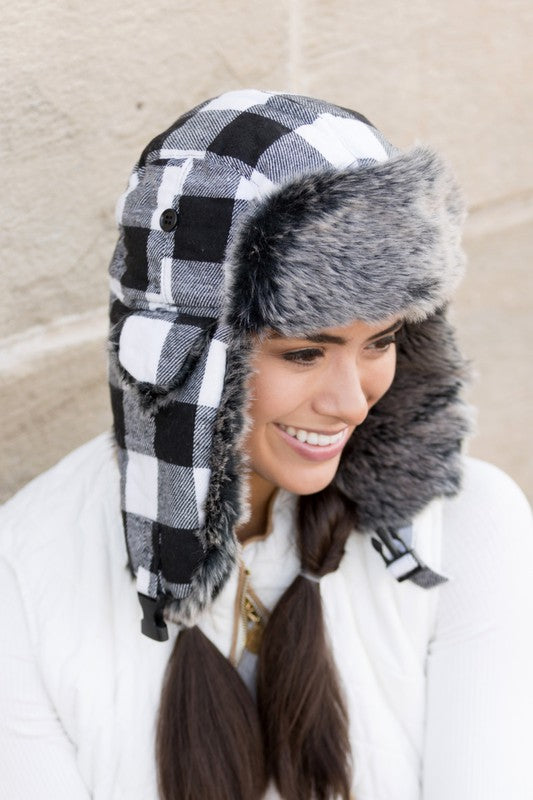 Woman smiling while wearing a checkered Buffalo Trapper Hat with ear flaps and faux fur interior.