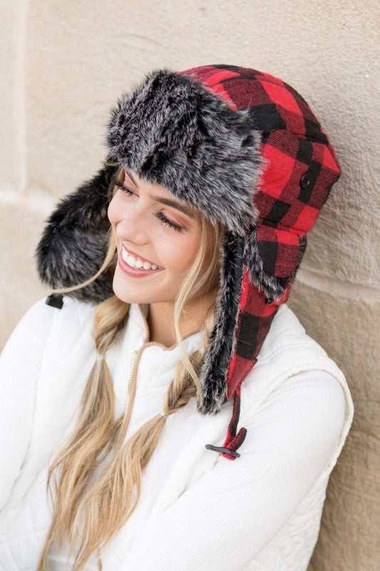 Woman smiling while wearing a checkered Buffalo Trapper Hat with ear flaps and faux fur interior.