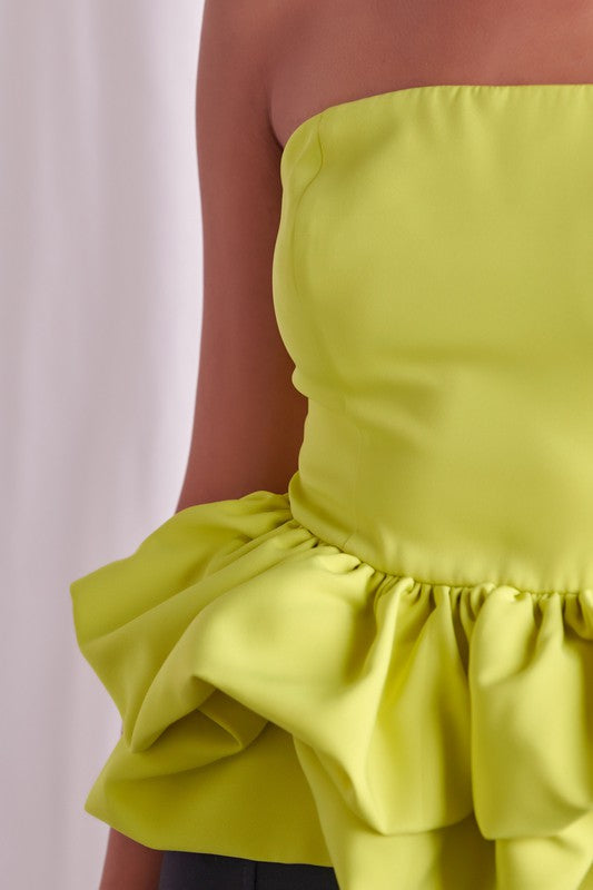 A woman stands against a light background, wearing an Off Shoulder Ruffle Top in bright pink and a shiny black skirt. She has long dark hair and is looking down while touching her hair with one hand.