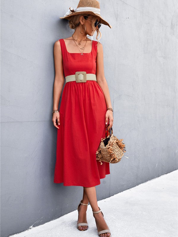 A woman in a Women's Sleeve Less Dress, featuring a vibrant red hue and crafted from 100% polyester, stands against a gray wall. She accessorizes with a wide-brimmed hat, holds a straw handbag, and complements her outfit with beige sandals.