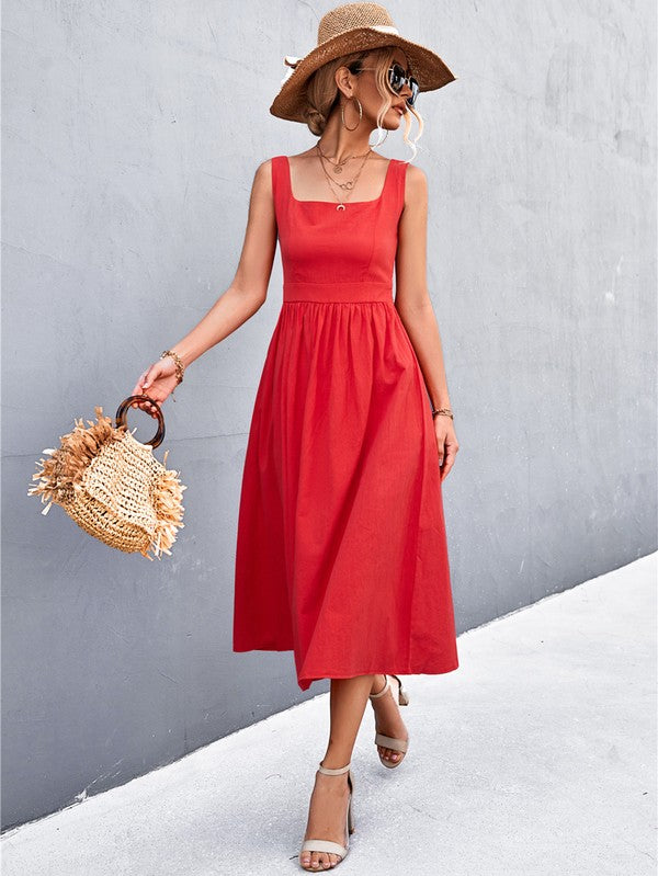 A woman in a Women's Sleeve Less Dress, featuring a vibrant red hue and crafted from 100% polyester, stands against a gray wall. She accessorizes with a wide-brimmed hat, holds a straw handbag, and complements her outfit with beige sandals.