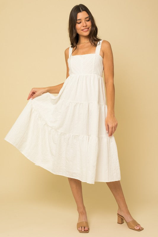 A woman in a white, sleeveless Textured Striped Smocked Back Midi Dress stands against a beige background, smiling softly. She pairs it with brown open-toe sandals.