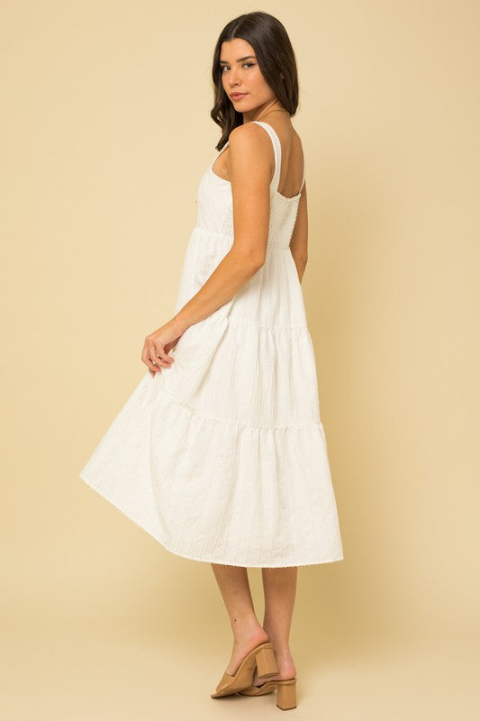 A woman in a white, sleeveless Textured Striped Smocked Back Midi Dress stands against a beige background, smiling softly. She pairs it with brown open-toe sandals.