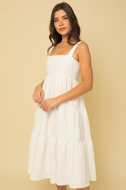 A woman in a white, sleeveless Textured Striped Smocked Back Midi Dress stands against a beige background, smiling softly. She pairs it with brown open-toe sandals.