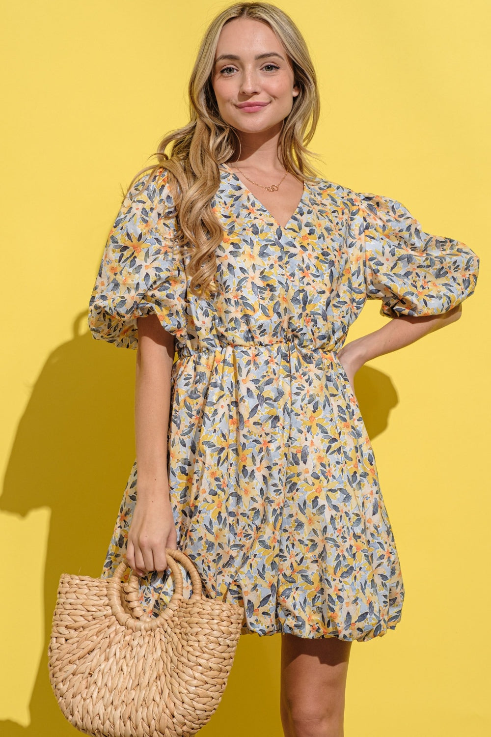 A person wearing the And The Why Full Size Floral Surplice Puff Sleeve Dress stands against a yellow background, holding a woven bag and wearing white sandals.