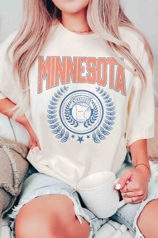 A woman sporting a 100% cotton MINNESOTA Graphic Tee with "Minnesota" emblazoned in bold orange letters and a decorative state emblem underneath, complemented by high-waisted denim shorts and a plaid shirt casually draped over her shoulders. The tee offers unisex sizing for an extra comfortable fit.