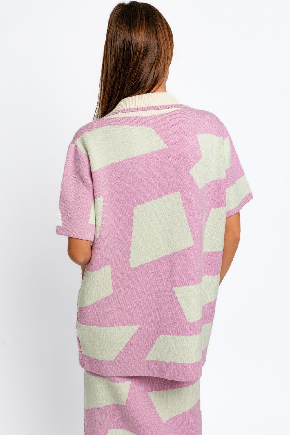 A woman is wearing the Le Lis Abstract Contrast Short Sleeve Collared Cardigan in pastel pink and cream with a geometric pattern, paired with a matching dress. She embodies a modern artistic touch as she looks to the side with her hands at her sides.