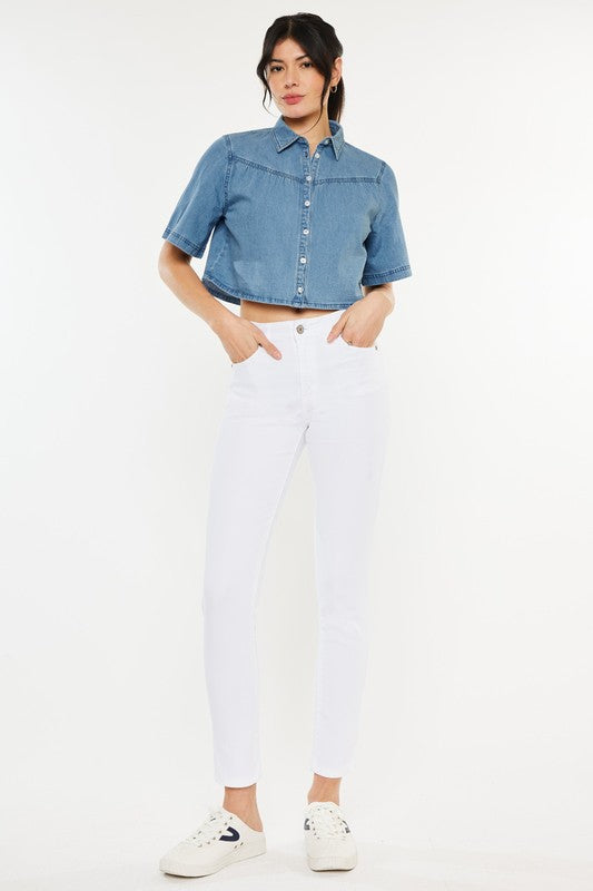 A person is wearing a denim short-sleeve button-up shirt, High Rise Ankle Skinny Jeans made with stretch fabric, and white sneakers, standing against a plain background.