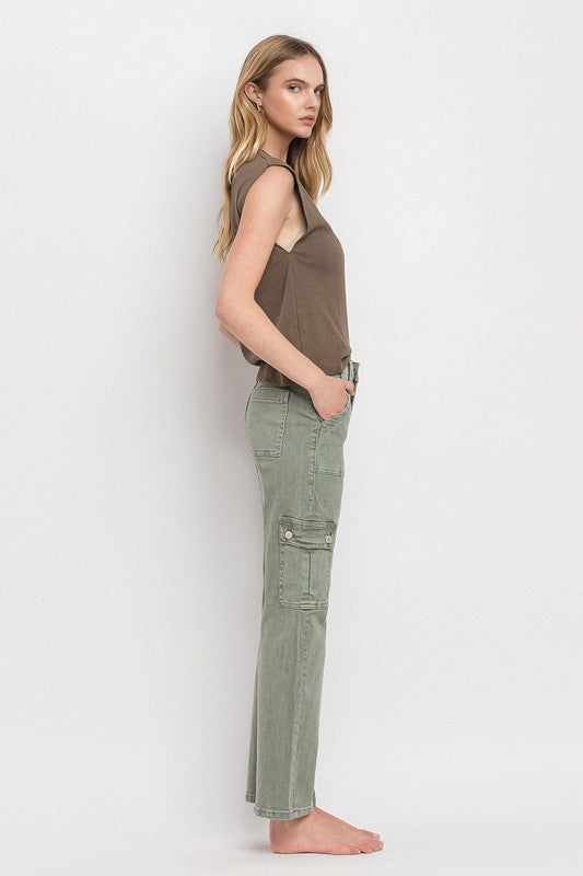 A person in high rise, relaxed-fit, army green cargo straight jeans and a brown shirt stands barefoot on a light background.