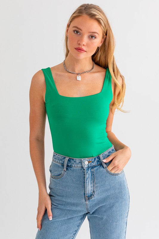 A person with long blonde hair wears the Square Neck Tank Bodysuit in green, perfectly styled for a comfortable fit, paired with blue jeans, while standing against a plain white background.