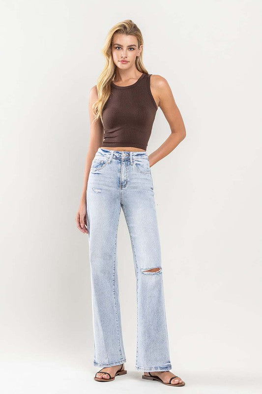 Person wearing light blue 90's Vintage Super High-Rise Flare Jeans with a large knee rip, paired with a brown sleeveless top and black sandals. The distressed denim adds an edgy touch to the ensemble.