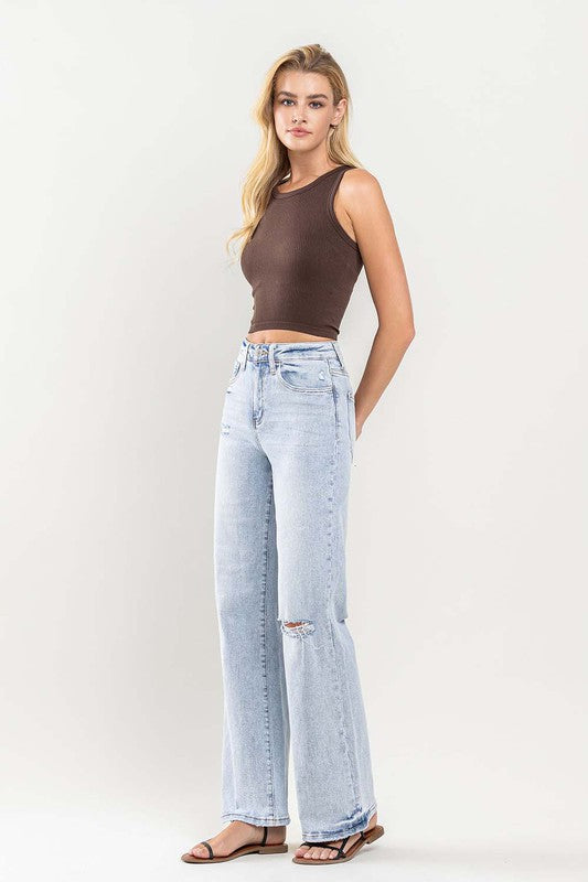 Person wearing light blue 90's Vintage Super High-Rise Flare Jeans with a large knee rip, paired with a brown sleeveless top and black sandals. The distressed denim adds an edgy touch to the ensemble.