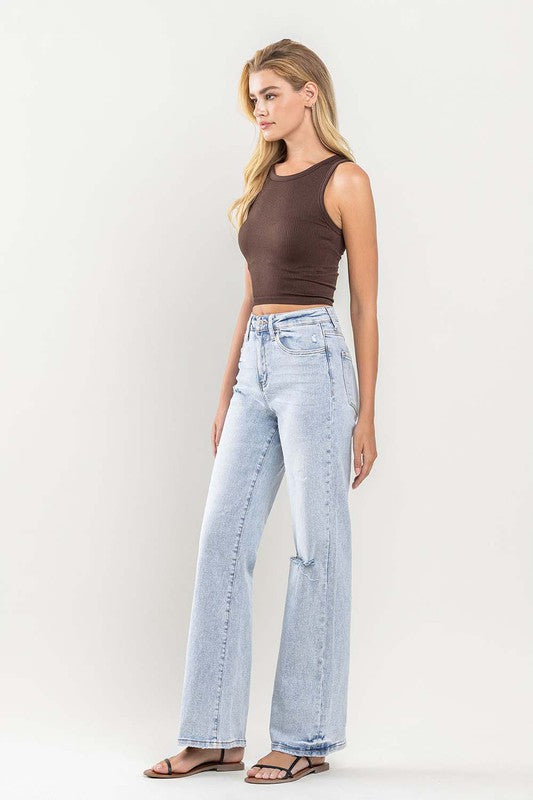 A person wearing light blue 90s Vintage Super High Rise Flare Jeans with a rip at the knee, paired with a brown top and sandals, stands against a neutral background.