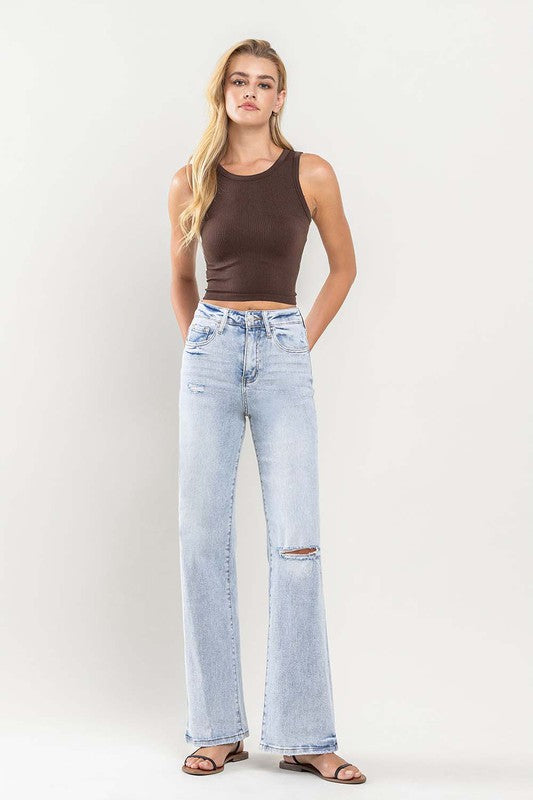 A person wearing light blue 90s Vintage Super High Rise Flare Jeans with a rip at the knee, paired with a brown top and sandals, stands against a neutral background.