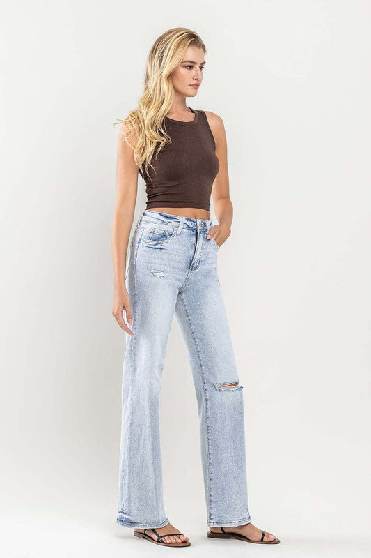 A person wearing light blue 90s Vintage Super High Rise Flare Jeans with a rip at the knee, paired with a brown top and sandals, stands against a neutral background.