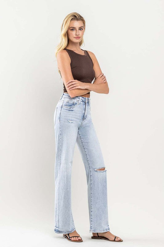 A person wearing light blue 90s Vintage Super High Rise Flare Jeans with a rip at the knee, paired with a brown top and sandals, stands against a neutral background.