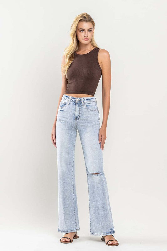 A person wearing light blue 90s Vintage Super High Rise Flare Jeans with a rip at the knee, paired with a brown top and sandals, stands against a neutral background.