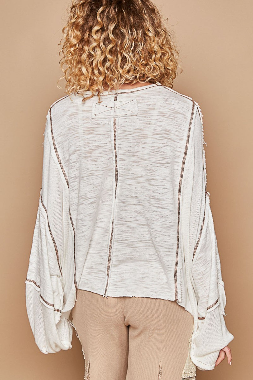 A person with curly shoulder-length hair wears a loose, white POL Round Neck Contrast Stitching High-Low Top and beige pants while posing against a beige background.