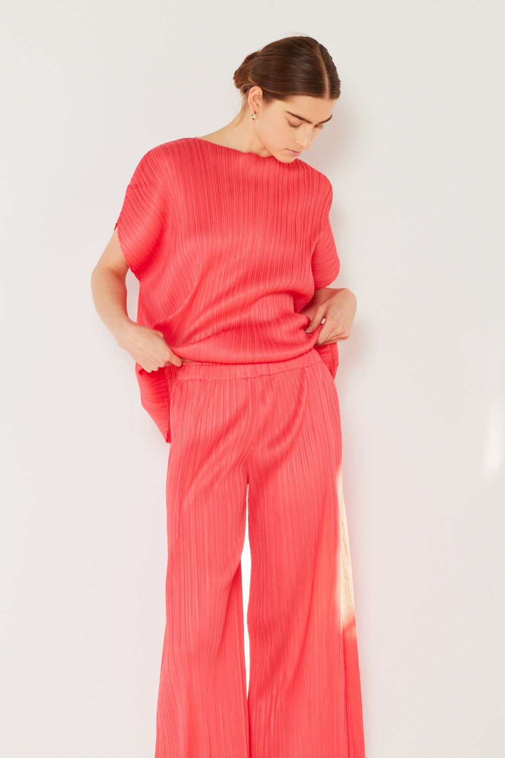 A person wearing the Marina West Swim Pleated Wide-Leg Pants with side pleat detail in red, standing against a white background with hands in pockets and wearing white sandals.