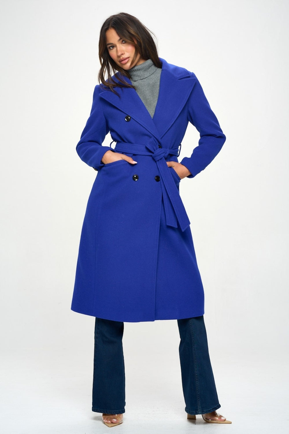 Person wearing the Coalition LA Double-Breasted Longline Coat with Belt, made from vegan wool in blue, over a grey turtleneck, posing against a plain background.