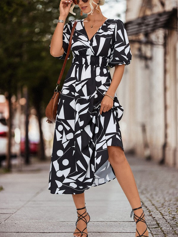 A person in a Women Printed Long Dress featuring a black and white geometric pattern and a V-neck stands outdoors, wearing strappy sandals and holding a brown handbag.