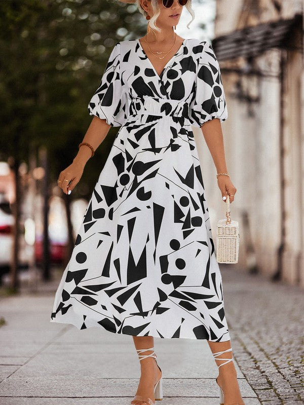 A person in a Women Printed Long Dress featuring a black and white geometric pattern and a V-neck stands outdoors, wearing strappy sandals and holding a brown handbag.