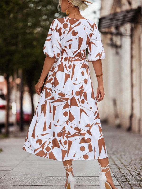 A person in a Women Printed Long Dress featuring a black and white geometric pattern and a V-neck stands outdoors, wearing strappy sandals and holding a brown handbag.