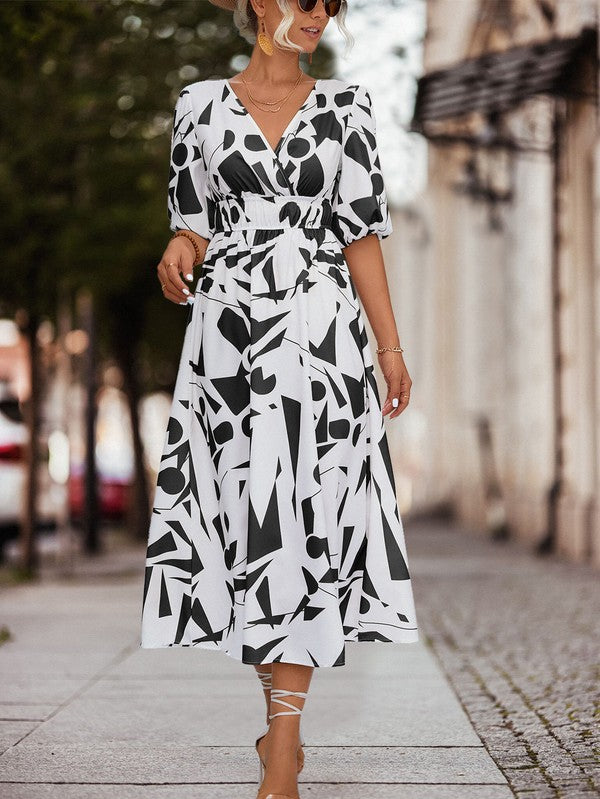 A person in a Women Printed Long Dress featuring a black and white geometric pattern and a V-neck stands outdoors, wearing strappy sandals and holding a brown handbag.