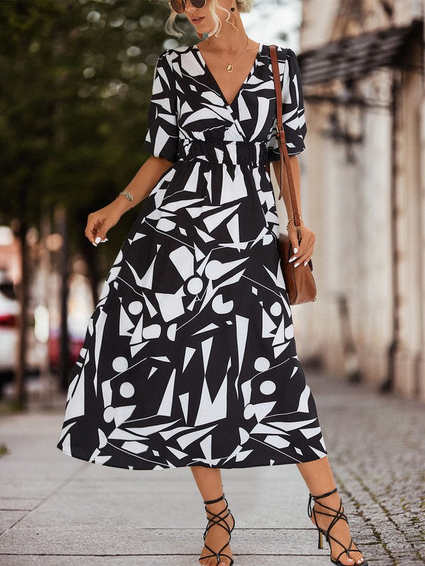 A person in a Women Printed Long Dress featuring a black and white geometric pattern and a V-neck stands outdoors, wearing strappy sandals and holding a brown handbag.