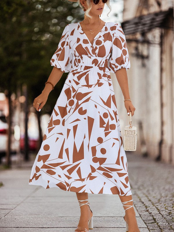 A person in a Women Printed Long Dress featuring a black and white geometric pattern and a V-neck stands outdoors, wearing strappy sandals and holding a brown handbag.