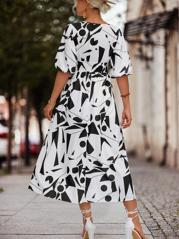 A person in a Women Printed Long Dress featuring a black and white geometric pattern and a V-neck stands outdoors, wearing strappy sandals and holding a brown handbag.