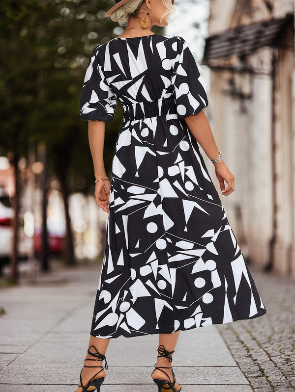 A person in a Women Printed Long Dress featuring a black and white geometric pattern and a V-neck stands outdoors, wearing strappy sandals and holding a brown handbag.